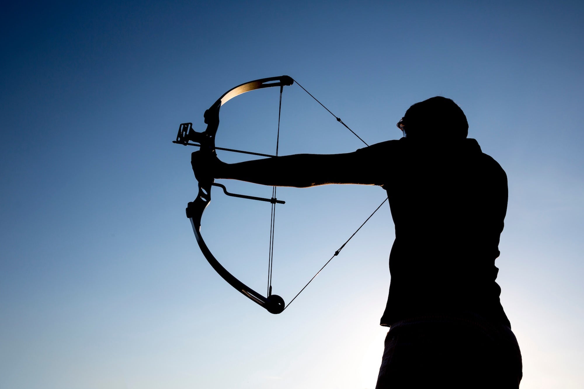 Archer silhouetted against the sky while drawing a compound bow during outdoor target practice.