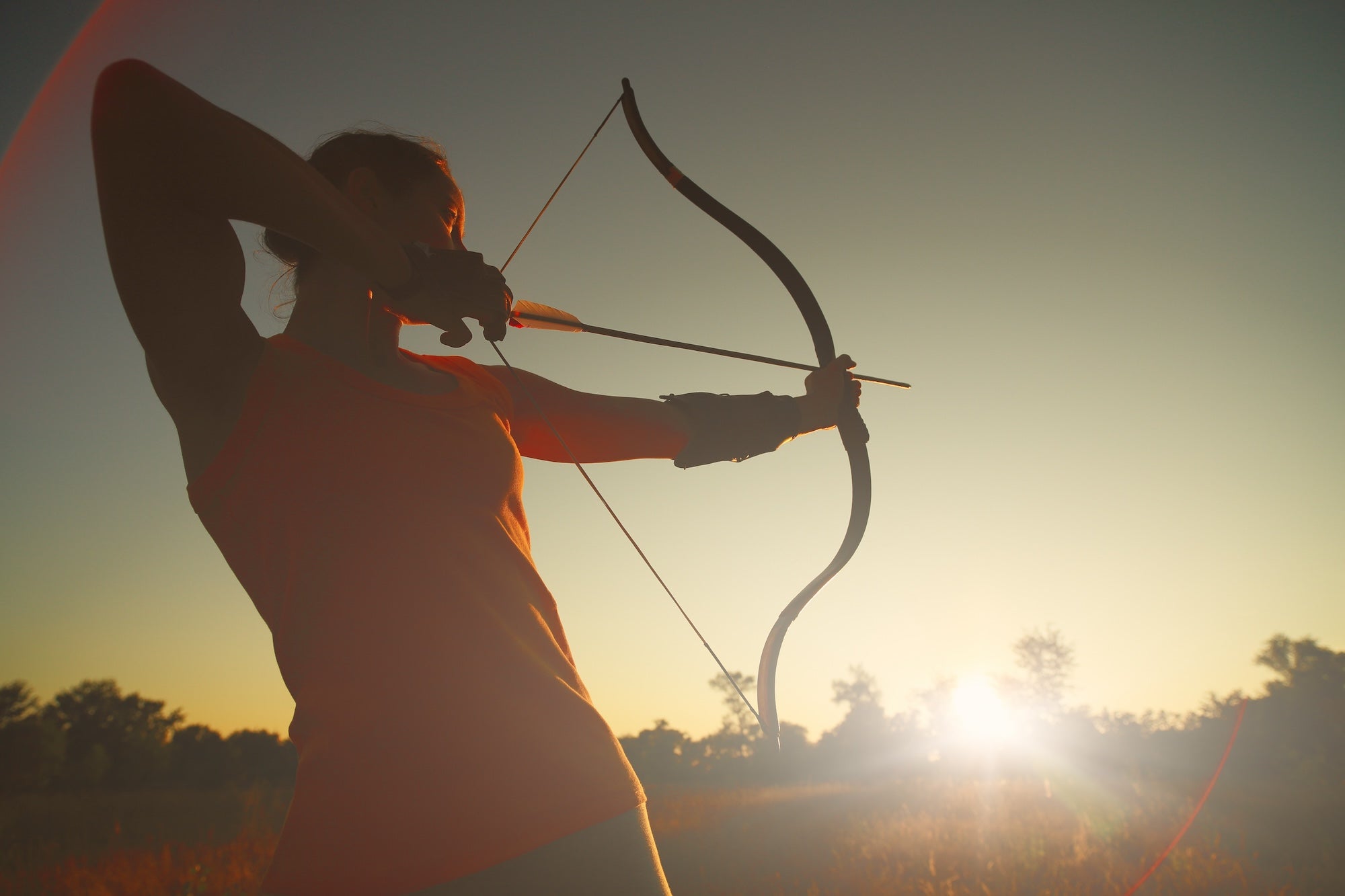 Archer aiming a Bear Archery bow at sunset in an open field, demonstrating focus and technique.